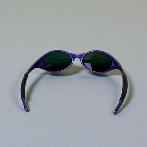 May include: Purple sunglasses with dark green lenses. The frames are a vibrant purple, and the arms have black rubber grips for comfort. The sunglasses are set against a neutral gray background.