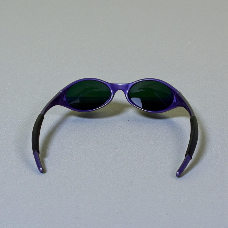 May include: Purple sunglasses with dark green lenses. The frames are a vibrant purple, and the arms have black rubber grips for comfort. The sunglasses are set against a neutral gray background.