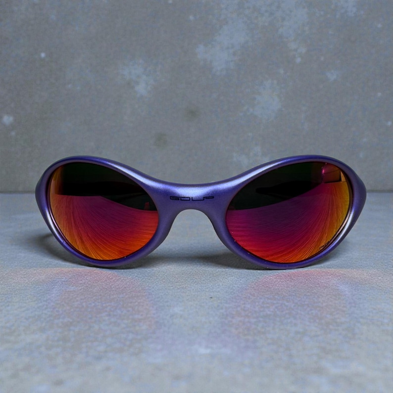 May include: Purple sunglasses with a curved frame and reflective red-orange lenses. The word "SOUP" is printed on the bridge of the sunglasses. The sunglasses are set against a gray background.
