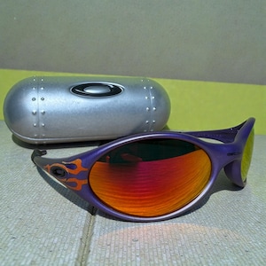 May include: Purple sunglasses with orange and red lenses and flame design accents. The sunglasses are next to a silver, oval-shaped case with the brand name "Oakley" on it. The lenses have a gradient effect.