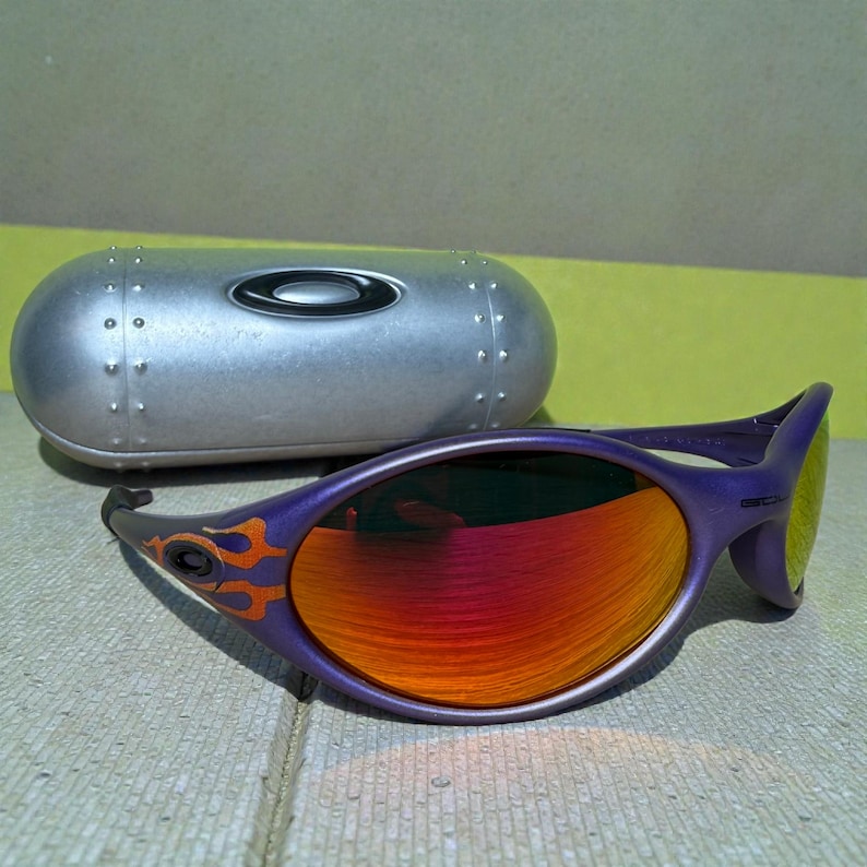 May include: Purple sunglasses with orange and red lenses and flame design accents. The sunglasses are next to a silver, oval-shaped case with the brand name "Oakley" on it. The lenses have a gradient effect.