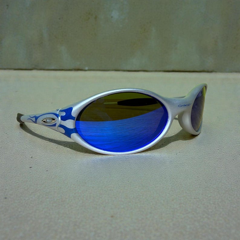 May include: Silver-framed sunglasses with blue mirrored lenses. The frame features a blue flame design on the temples. The lenses reflect a vibrant blue hue, and the overall design is sporty and modern.