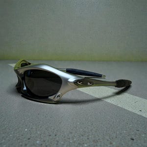 May include: Silver and black sunglasses with a sleek, modern design. The sunglasses feature a curved frame, dark lenses, and a black arm with a rubberized grip. The lenses are dark, and the frame has a metallic finish.