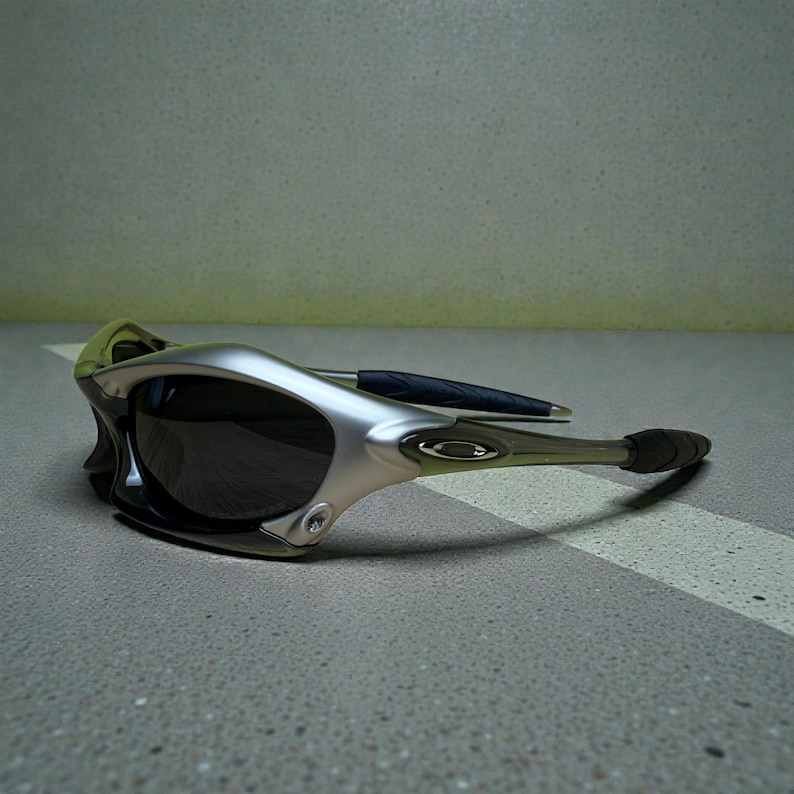 May include: Silver and black sunglasses with a sleek, modern design. The sunglasses feature a curved frame, dark lenses, and a black arm with a rubberized grip. The lenses are dark, and the frame has a metallic finish.
