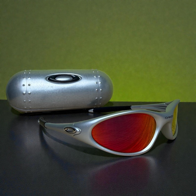 Puede incluir: Gafas de sol plateadas con lentes rojas y estuche plateado. Las gafas tienen una montura curvada y el logotipo de la marca en la patilla. El estuche es de forma ovalada con un acabado met&aacute;lico.