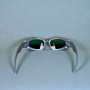 May include: Silver-framed sunglasses with green lenses. The sunglasses have a sleek, modern design with curved temples. The lenses reflect a pink hue. The overall aesthetic is sporty and stylish, suitable for outdoor activities.