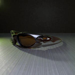 May include: Dark gray and brown sunglasses with a wraparound design. The sunglasses have a metallic finish and brown tinted lenses. The frame features a unique, angular shape with a small logo on the side. The sunglasses are designed for sports and outdoor activities.