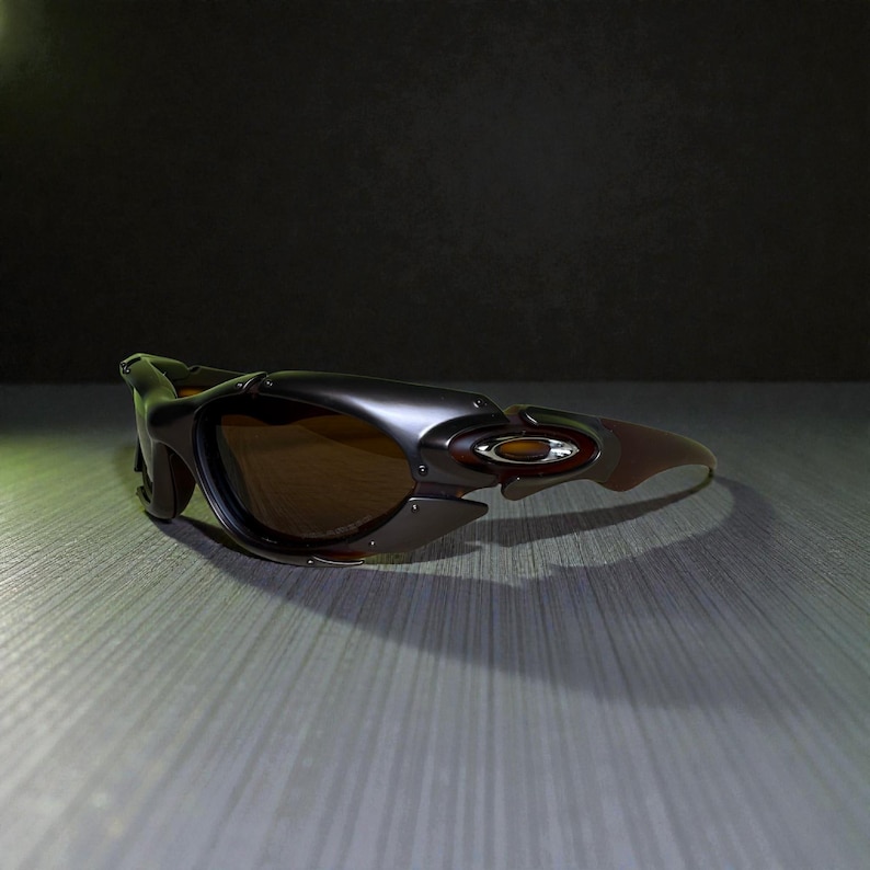 May include: Dark gray and brown sunglasses with a wraparound design. The sunglasses have a metallic finish and brown tinted lenses. The frame features a unique, angular shape with a small logo on the side. The sunglasses are designed for sports and outdoor activities.
