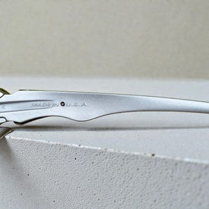 May include: Close-up of a silver metal object with the text "MADE IN U.S.A." engraved on its surface. The object has a curved, elongated shape with a decorative edge. The background is a light, neutral color.
