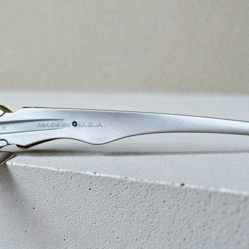 May include: Close-up of a silver metal object with the text "MADE IN U.S.A." engraved on its surface. The object has a curved, elongated shape with a decorative edge. The background is a light, neutral color.
