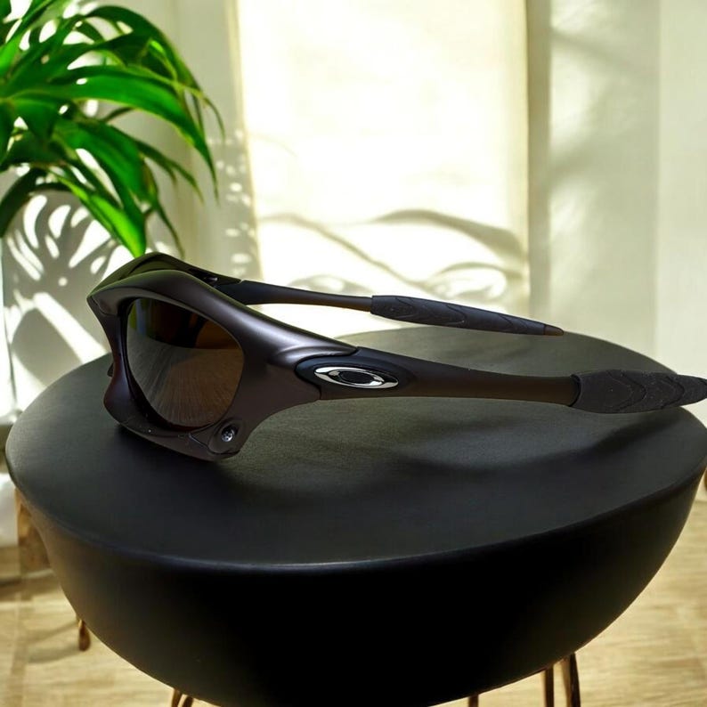 May include: Dark brown sunglasses with a wraparound design. The sunglasses have a curved frame and dark lenses. The arms of the sunglasses are black with textured grips. The Oakley logo is visible on the frame.