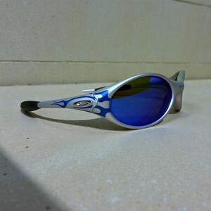 May include: Silver sunglasses with blue mirrored lenses and a flame design in blue. The sunglasses have a sleek, modern design with a black temple tip. The lenses are oval-shaped.