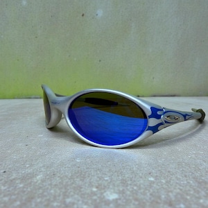 May include: Silver-framed sunglasses with blue mirrored lenses. The temples feature a blue flame design. The lenses reflect a blue hue. The sunglasses are set against a neutral background.