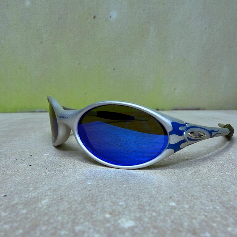 May include: Silver-framed sunglasses with blue mirrored lenses. The temples feature a blue flame design. The lenses reflect a blue hue. The sunglasses are set against a neutral background.