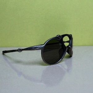 May include: Dark grey sunglasses with a rounded frame and dark lenses. The sunglasses have a sleek, modern design with a curved temple and a small logo on the side. The lenses are dark, providing shade from the sun.