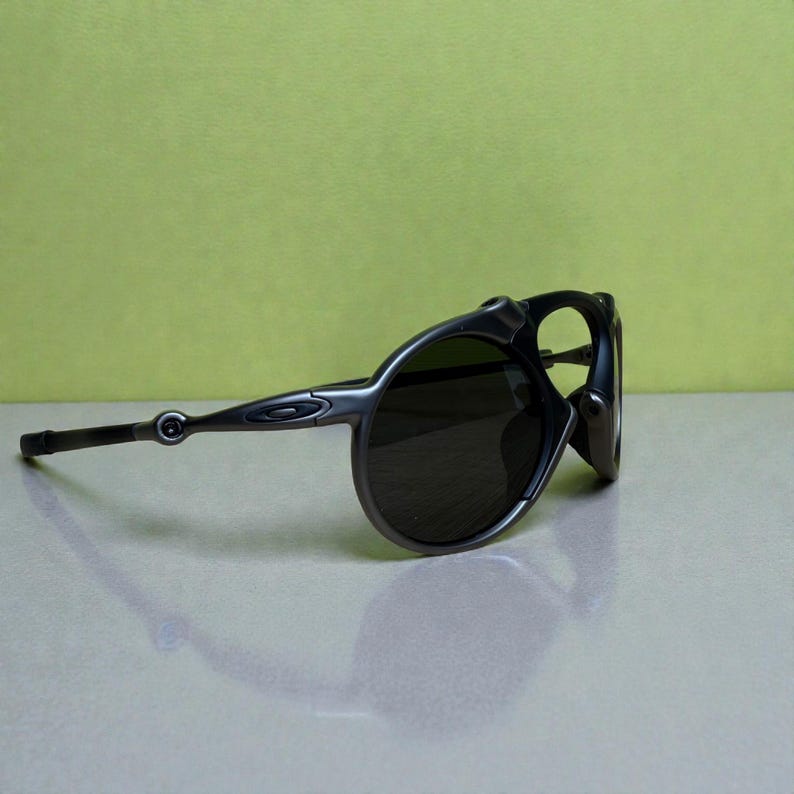 May include: Dark grey sunglasses with a rounded frame and dark lenses. The sunglasses have a sleek, modern design with a curved temple and a small logo on the side. The lenses are dark, providing shade from the sun.