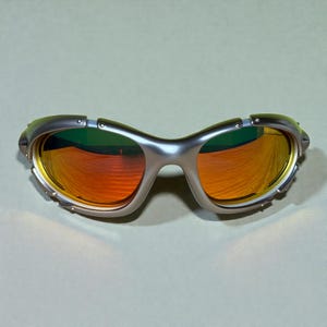 May include: Silver-framed sunglasses with orange and yellow lenses. The sunglasses have a sporty design with a curved frame and visible hinges. The lenses reflect a gradient of colors, and the frame has a metallic finish.