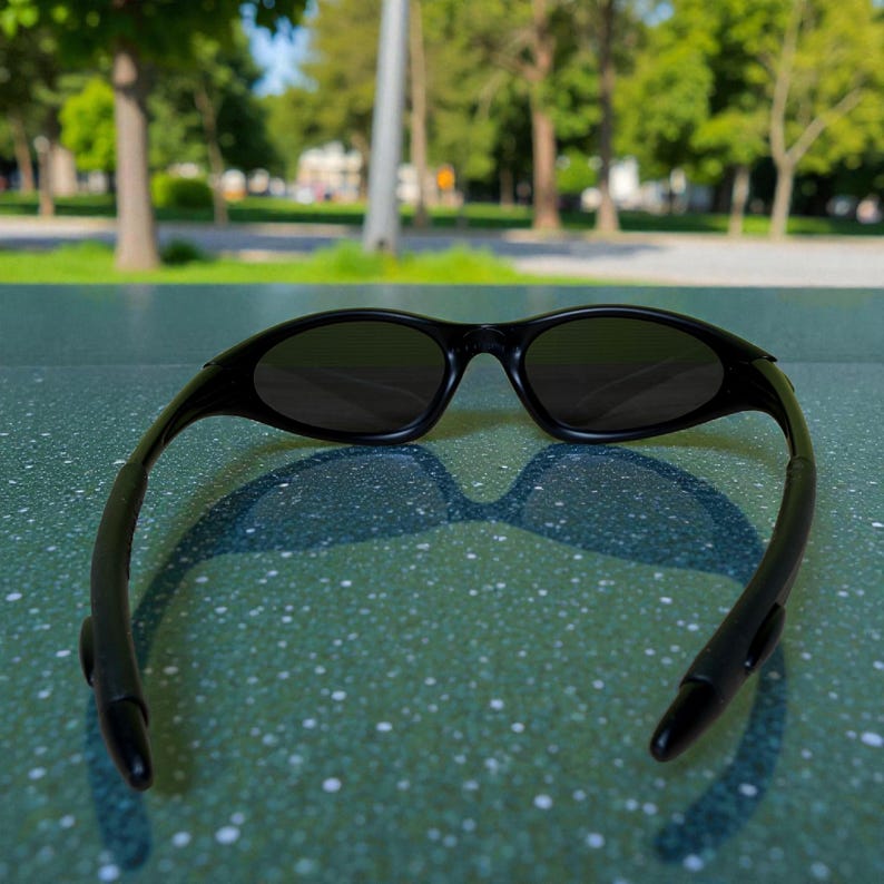 K&ouml;nnte beinhalten: Schwarze Sonnenbrille mit dunklen Gl&auml;sern. Die Sonnenbrille liegt auf einer gr&uuml;n gesprenkelten Oberfl&auml;che, mit einem verschwommenen Hintergrund aus B&auml;umen und einem Park. Die Sonnenbrille hat einen sportlichen Stil.