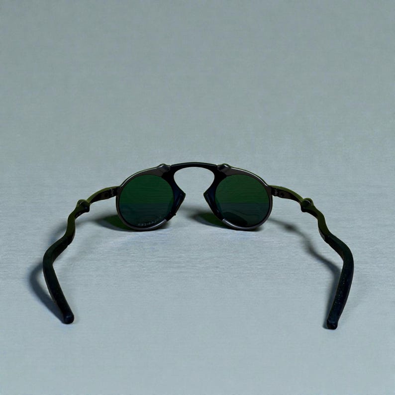 May include: Black sunglasses with round green lenses. The frame is black with a unique, curved design on the temples. The sunglasses are set against a light gray background.