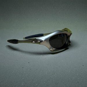 May include: Silver and olive green sunglasses with dark lenses. The sunglasses have a sleek, modern design with a curved frame and black accents on the arms. The Oakley logo is visible on the side of the frame.