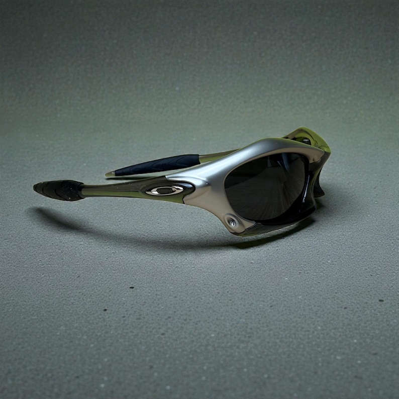 May include: Silver and olive green sunglasses with dark lenses. The sunglasses have a sleek, modern design with a curved frame and black accents on the arms. The Oakley logo is visible on the side of the frame.