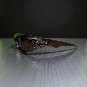 May include: A pair of Oakley sunglasses with a brown and black frame and tinted lenses. The sunglasses have a sleek, modern design with a curved shape and a metallic accent on the side. The lenses are a gradient brown color.