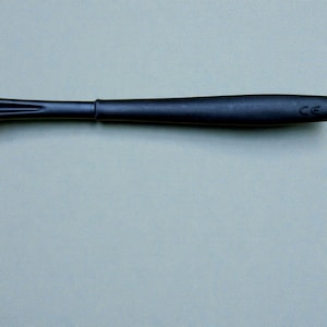 K&ouml;nnte beinhalten: Nahaufnahme eines schwarzen Utensils, m&ouml;glicherweise eines K&uuml;chenwerkzeugs. Das Objekt hat einen langen, schlanken Griff und ein breiteres, abgerundetes Ende. Die Oberfl&auml;che erscheint glatt und matt, mit subtilen Details.