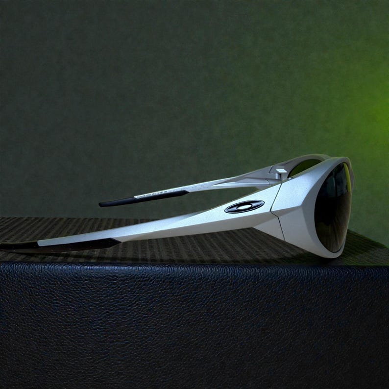 Op de afbeelding: Zilverkleurige en zwarte sportzonnebril met een strak, modern design. De zonnebril heeft een wrap-around stijl met donkere glazen en een zilverkleurig montuur. Het Oakley-logo is zichtbaar aan de zijkant. De zonnebril ligt op een getextureerd oppervlak.