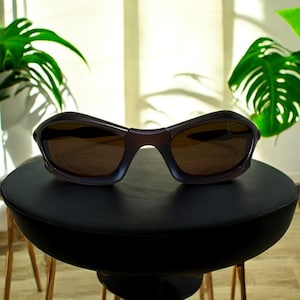 May include: Dark brown sunglasses with a curved frame and tinted lenses. The sunglasses are resting on a small, round, black table. The table has three gold-colored legs. Green plants are in the background.