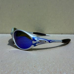May include: Silver sunglasses with blue mirrored lenses and a flame design in blue. The arms have black tips and the frame is silver. The sunglasses are angled slightly, showcasing the reflective lenses.