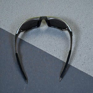 May include: Sunglasses with a black frame and dark lenses. The sunglasses have a silver accent on the top of the frame and black rubber grips on the arms. The lenses are rectangular and the overall design is sporty.