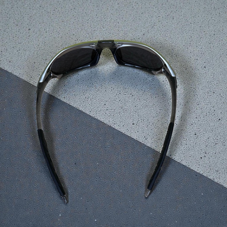 May include: Sunglasses with a black frame and dark lenses. The sunglasses have a silver accent on the top of the frame and black rubber grips on the arms. The lenses are rectangular and the overall design is sporty.