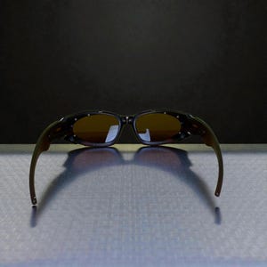 May include: Sunglasses with a dark frame and brown lenses. The sunglasses have a curved design with a dark brown and olive green color scheme. The lenses are a light brown color.
