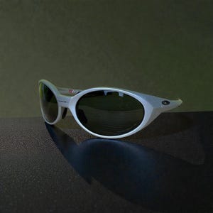 Op de afbeelding: Witte zonnebril met donkere glazen. De zonnebril heeft een uniek, hoekig montuurontwerp. Het Oakley-logo is zichtbaar op de poot. De zonnebril staat tegen een donkere achtergrond.