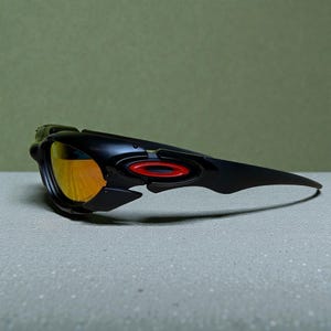 Może przedstawiać: Czarne okulary przeciwsłoneczne z pomarańczowymi szkłami. Okulary mają elegancki, nowoczesny design z czerwonym akcentem z boku. Oprawka jest matowa czarna, a soczewki odbijają złoty odcień.