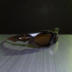 May include: A pair of black and brown sunglasses with a sporty design. The sunglasses have brown lenses and a dark frame with a red and gold logo on the side. The arms are a reddish-brown color.