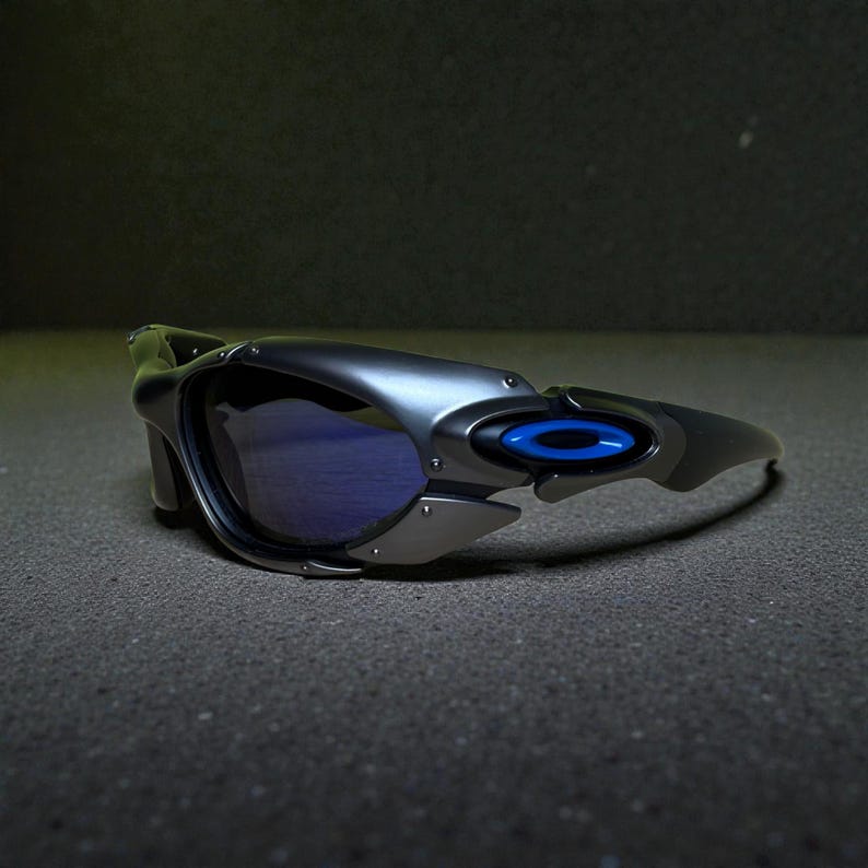 May include: Sleek, metallic gray sunglasses with blue lenses. The sunglasses feature a modern, angular design with a blue accent on the temple. The lenses are a deep blue, offering a contrast to the gray frame.