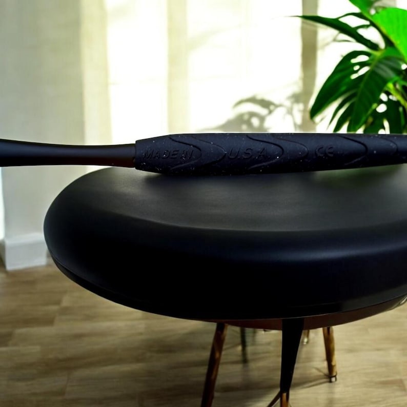 May include: A black baseball bat with a textured grip, resting on a black stool. The grip is embossed with the words "MADE IN USA". The bat has a dark brown barrel and is set against a blurred background.