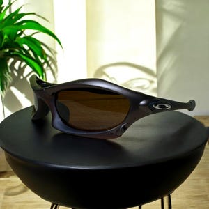 May include: Dark brown sunglasses with a curved, wraparound design. The lenses are a warm brown color, and the frames have a sleek, modern aesthetic. The sunglasses are resting on a black surface.