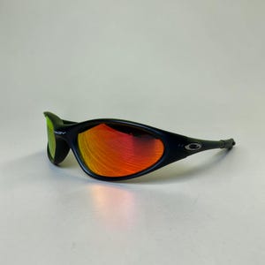 May include: Black sunglasses with a curved frame and red-orange lenses. The lenses reflect light, creating a gradient effect. The sunglasses have a sleek, modern design, suitable for outdoor activities.