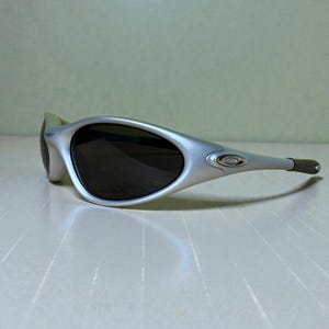 K&ouml;nnte beinhalten: Silberfarbene Sonnenbrille mit dunklen Gl&auml;sern. Die Sonnenbrille hat ein geschwungenes Design mit einem silberfarbenen Rahmen und dunklen Gl&auml;sern. Die B&uuml;gel sind dunkelgrau. Das Oakley-Logo ist auf der Seite sichtbar.