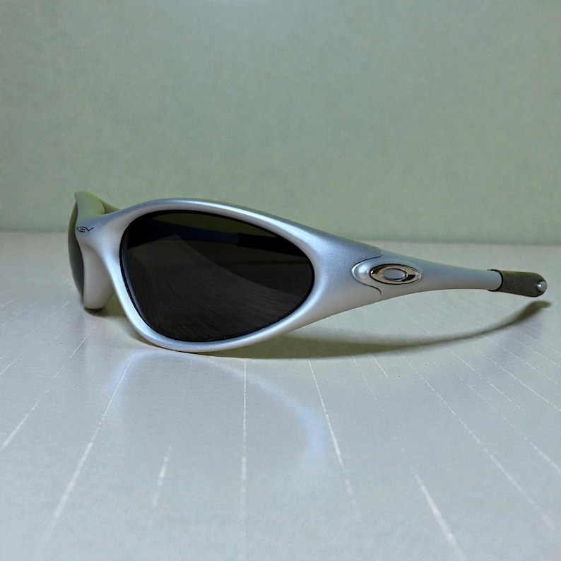 K&ouml;nnte beinhalten: Silberfarbene Sonnenbrille mit dunklen Gl&auml;sern. Die Sonnenbrille hat ein geschwungenes Design mit einem silberfarbenen Rahmen und dunklen Gl&auml;sern. Die B&uuml;gel sind dunkelgrau. Das Oakley-Logo ist auf der Seite sichtbar.