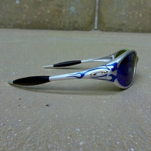May include: Silver sunglasses with blue flame design on the arms and dark blue lenses. The sunglasses have black earpieces and a sleek, modern design. The lenses appear to have a mirrored finish.