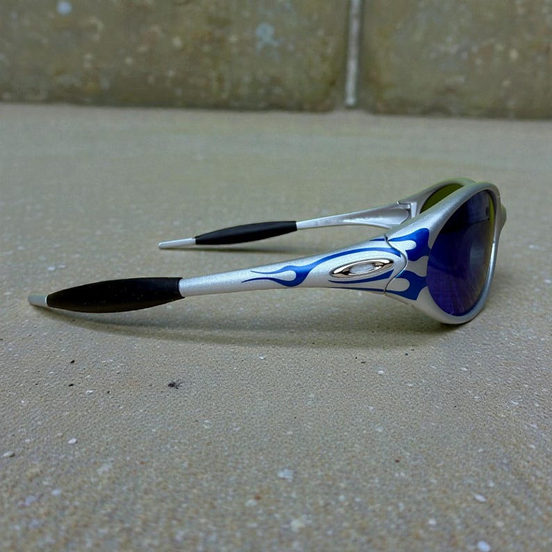 May include: Silver sunglasses with blue flame design on the arms and dark blue lenses. The sunglasses have black earpieces and a sleek, modern design. The lenses appear to have a mirrored finish.