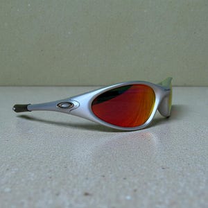 Op de afbeelding: Zilverkleurige zonnebril met rood-oranje glazen. De zonnebril heeft een gestroomlijnd ontwerp met een gebogen montuur en een klein logo op de poot. De glazen reflecteren een kleurengradi&euml;nt.