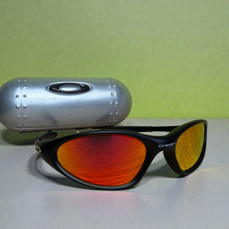 May include: Black Oakley sunglasses with red and yellow mirrored lenses. The sunglasses have a sleek, curved design and are accompanied by a silver case. The Oakley logo is visible on the frame.