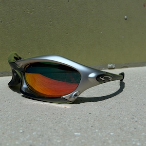 May include: Silver and black sunglasses with a mirrored lens that reflects orange and red hues. The sunglasses have a sleek, modern design with a curved frame and a small logo on the temple.