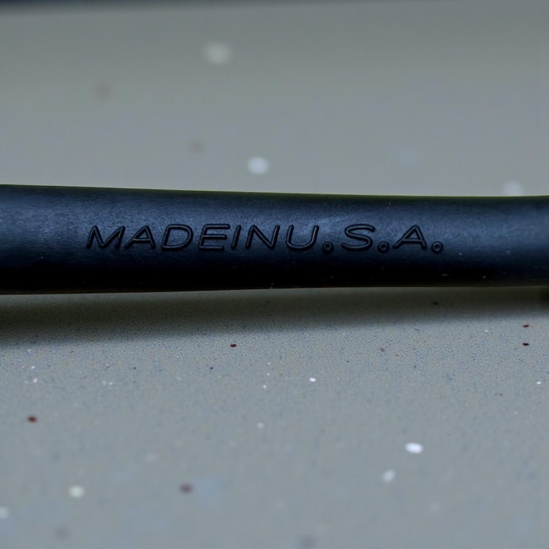 Op de afbeelding: Close-up van een zwart object met de in reli&euml;f aangebrachte tekst "MADE IN U.S.A.". Het object lijkt een handvat of onderdeel van een gereedschap te zijn, met een matte afwerking. De achtergrond is een lichtgrijze ondergrond.