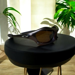 May include: Brown and black sunglasses with a curved frame and tinted lenses. The sunglasses are resting on a black surface. The Oakley logo is visible on the side of the frame. The sunglasses are designed for sports and outdoor activities.
