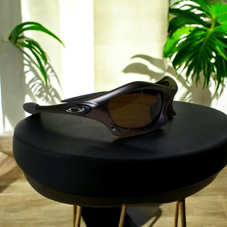 May include: Brown and black sunglasses with a curved frame and tinted lenses. The sunglasses are resting on a black surface. The Oakley logo is visible on the side of the frame. The sunglasses are designed for sports and outdoor activities.